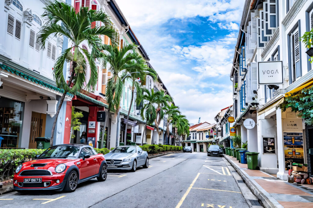A bright street view in the Tiong Bahru neighborhood, lined with colorful, preserved shophouses and palm trees, with vintage and luxury cars parked on the street.