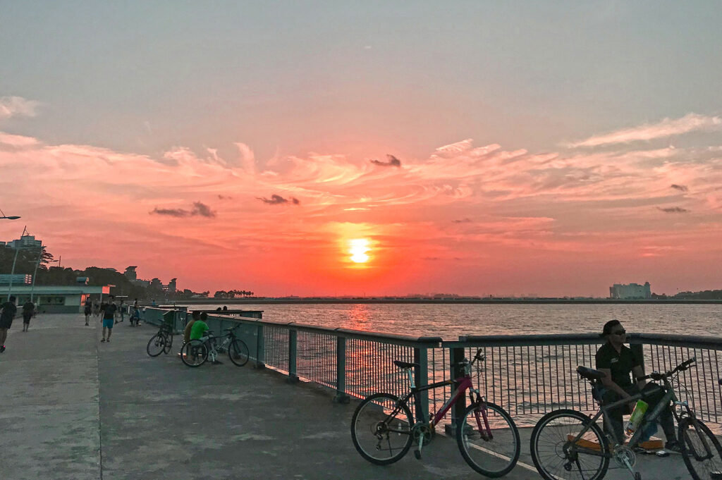 Warm, vibrant sunset over a calm body of water, viewed from a waterfront promenade with people and parked bicycles silhouetted against the bright orange and pink sky.