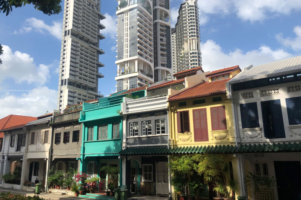 Vibrant scene showing a row of beautifully restored, brightly colored traditional Singapore shophouses (teal, yellow, gray) standing in sharp contrast beneath towering, modern high-rise condominiums.