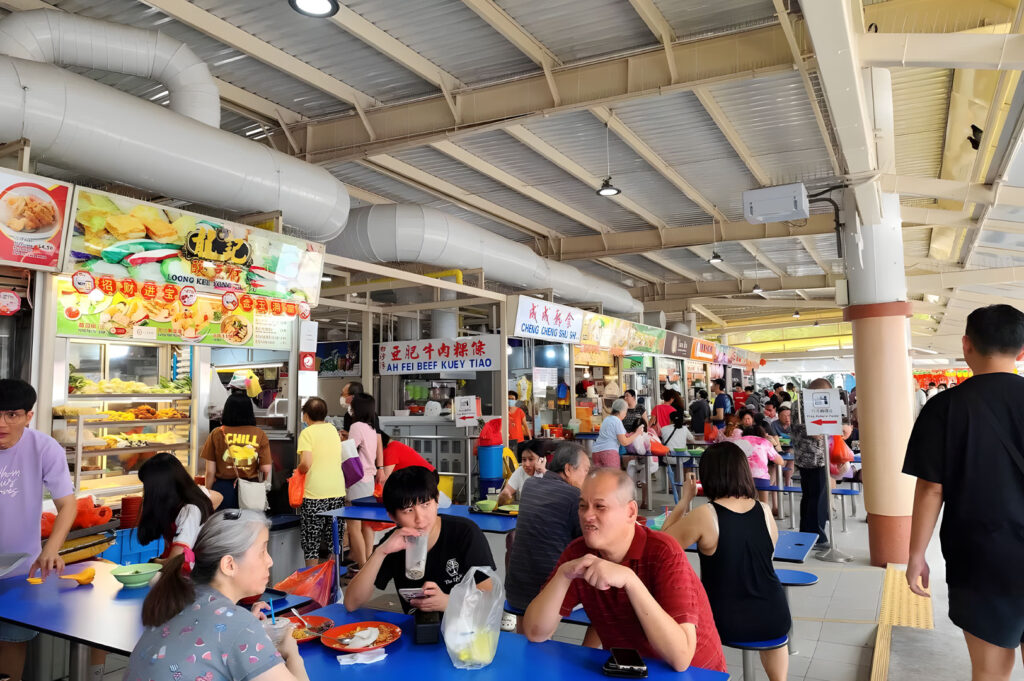Busy, brightly lit outdoor or covered hawker center with vendors and many people seated at blue rectangular tables, enjoying meals in a vibrant, communal setting.