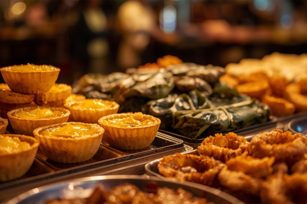 A close-up view of a selection of festive foods, including small tarts with yellow filling, items wrapped in leaves, and fried savory snacks.