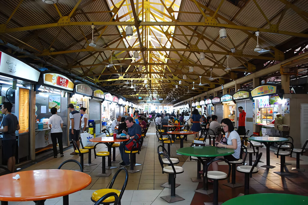 A long, wide-angle interior view of the famous Maxwell Food Centre in Singapore, showing the open, industrial roof structure and rows of colorful hawker stalls with round tables and chairs.