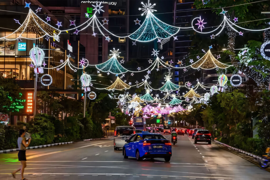 A night scene of cars driving along Orchard Road in Singapore, adorned with elaborate, glittering Christmas or festive light decorations strung across the street.