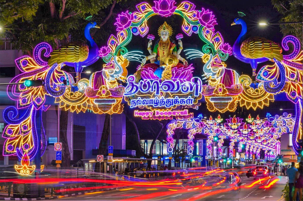 Night photo of the spectacular Deepavali (Diwali) light-up archway in Little India, featuring a brightly illuminated arch of the Goddess Lakshmi flanked by two peacocks, with streetlights blurred below.