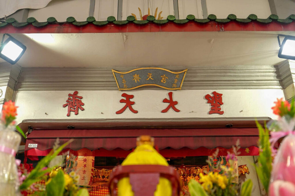 Close-up of the entrance signage of a Chinese temple (possibly the Qi Tian Gong or Monkey God Temple in Tiong Bahru) with red and gold characters on a white wall, framed by flowers and modern lights.