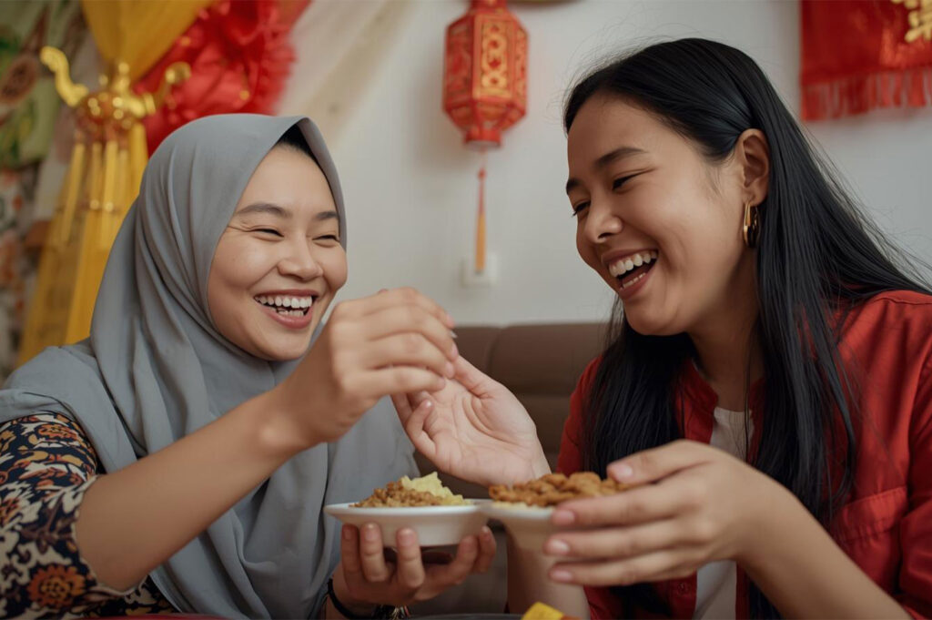 Two women, one wearing a gray hijab, laughing while sharing small plates of food in a celebratory setting with Chinese lanterns in the background.