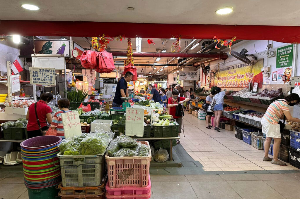 A wide-angle view inside a bright heartland wet market (likely at Rivervale Plaza) in Singapore, showing vendors selling fresh leafy greens and vegetables to local residents.