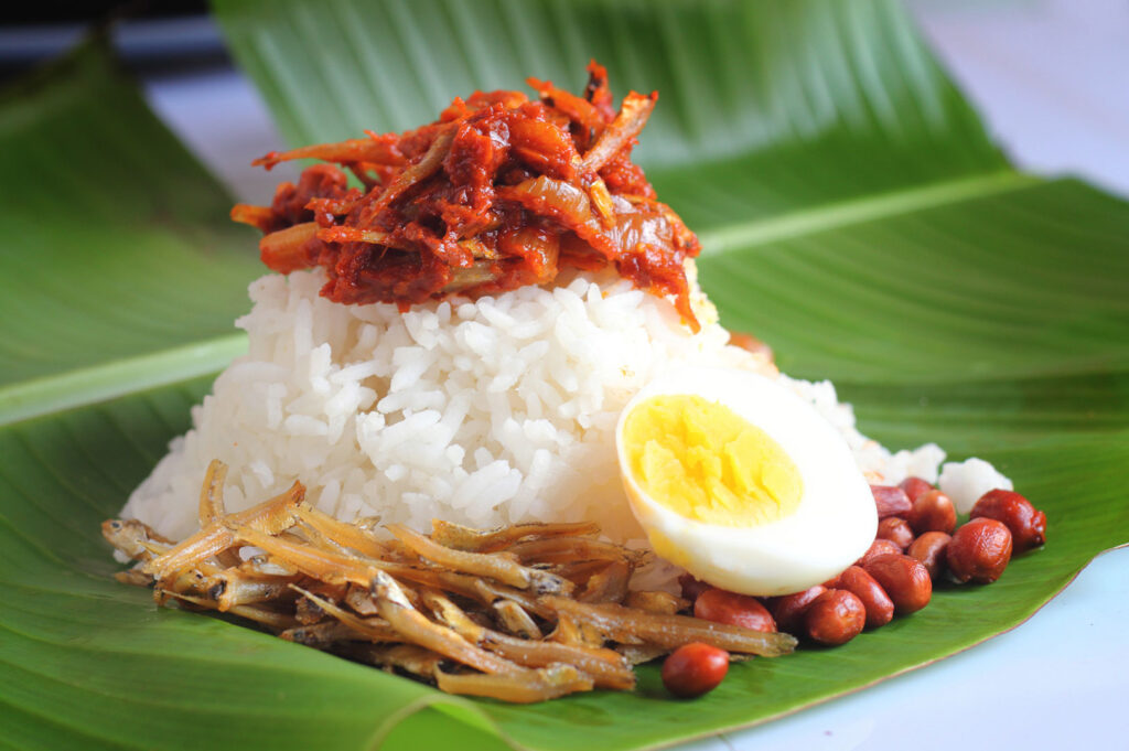 Close-up of Nasi Lemak served on a banana leaf: a mound of coconut rice topped with bright red sambal anchovies (ikan bilis), roasted peanuts, fried anchovies, and a hard-boiled egg.