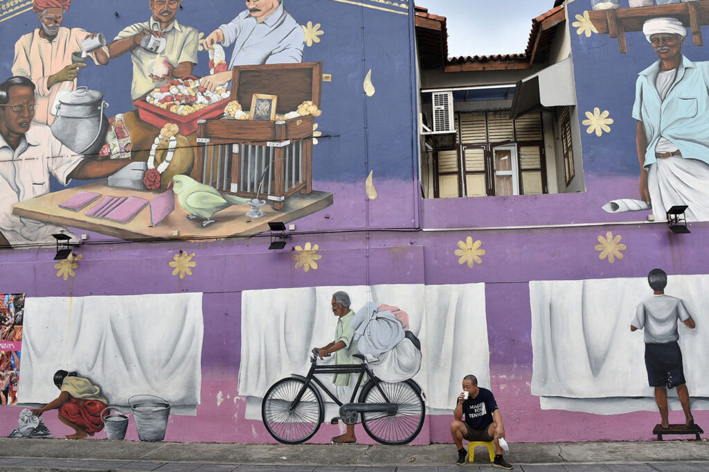 Large, colorful two-story street art mural in Little India depicting scenes of local life, including men making drinks, a cyclist, and people washing clothes, painted on a purple wall.