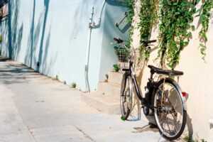 A black bicycle with a silver wire basket rests against a cream-colored wall draped with lush green ivy near a small set of concrete steps. Sunlight casts distinct tree shadows across the adjacent light blue wall, highlighting the quiet, sunny urban atmosphere.