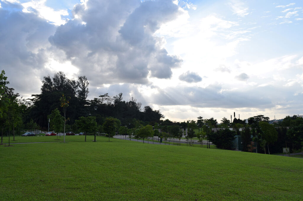 Wide landscape view of a large, manicured grassy slope in an open park area, with sun rays breaking through dramatic clouds over a distant tree line.