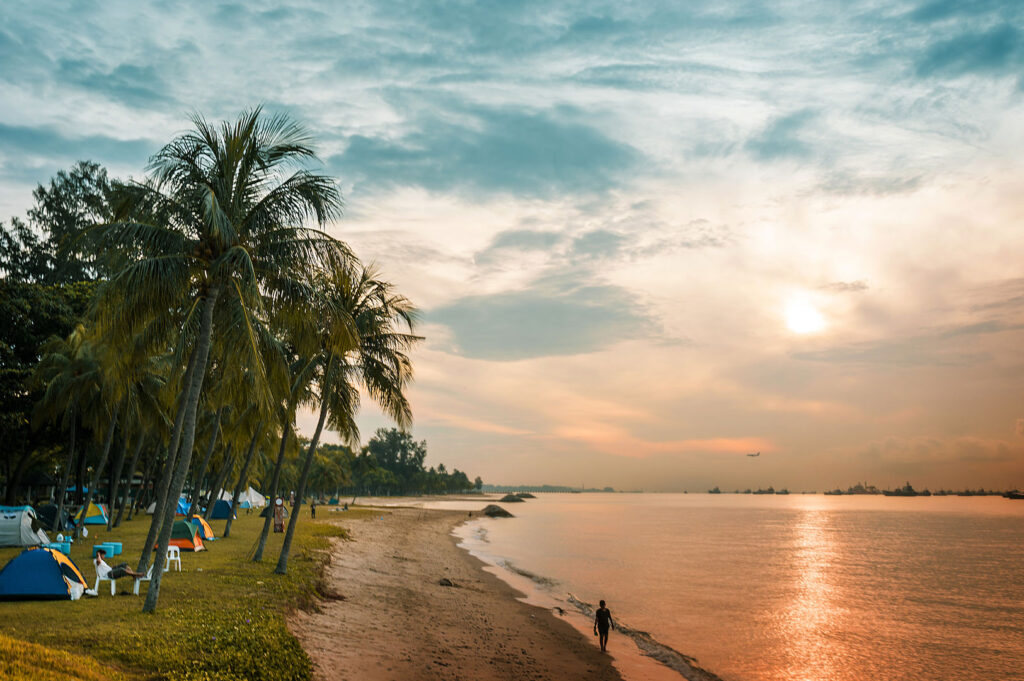 Scenic sunrise or sunset view over a beach, with palm trees and a row of colorful tents and campers along the sand, and a lone figure walking along the water's edge.