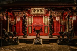 This ornate traditional Chinese temple features vibrant red doors and intricate pillars wrapped in sculpted dragons, surmounted by a golden plaque with Chinese calligraphy. In the foreground, weathered stone guardian statues and mythical creatures flank a central incense burner, creating a solemn and historic atmosphere.