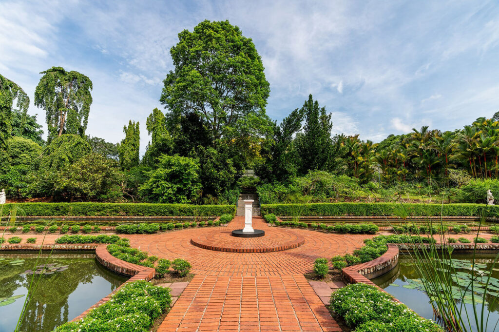 Wide view of a formal garden featuring a semi-circular red brick path leading to a white pedestal statue, surrounded by a pond and lush, towering tropical foliage.