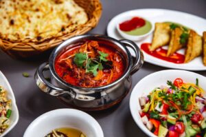 Close-up of a metal bowl with rich red Indian curry, garnished with cilantro, served with naan bread and samosas on a table in Singapore.