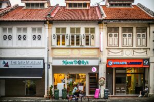 This image displays a colorful row of traditional shophouses featuring distinct businesses, including The Gem Museum and a laundry service. At street level, a cyclist rides past the storefronts while another person sits leisurely outside the shop on the far right.