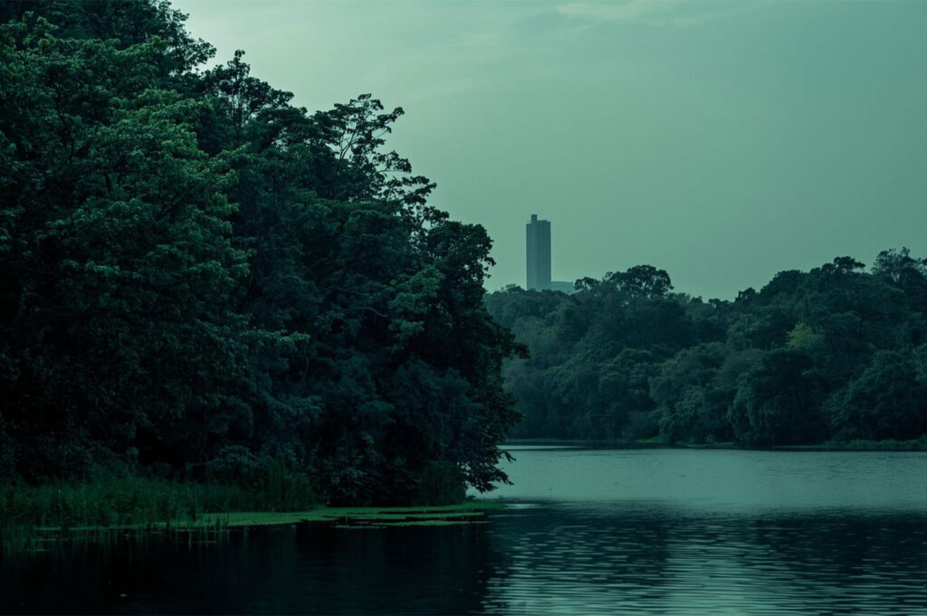Moody, twilight-toned photo of a dark, forested shoreline reflected in a still lake, with a single modern high-rise building visible in the distant background.