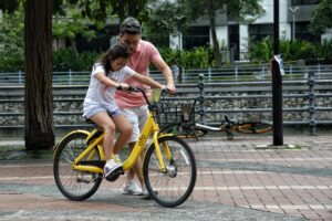 A man in a pink shirt assists a young girl as she learns to ride a bright yellow bicycle on a paved path. He walks closely alongside her, holding the handlebars to help steady the bike while she focuses on pedaling.