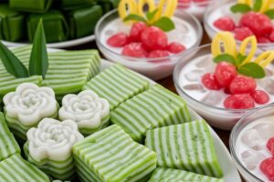 This close-up image showcases a variety of traditional Southeast Asian desserts, featuring distinct white and green striped layered cakes and flower-shaped gelatinous sweets in the foreground.