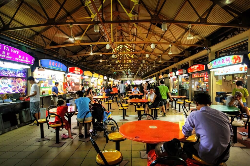 Bustling interior view of a Singaporean hawker center, showing rows of food stalls, brightly colored signs, and people seated at orange and yellow tables.