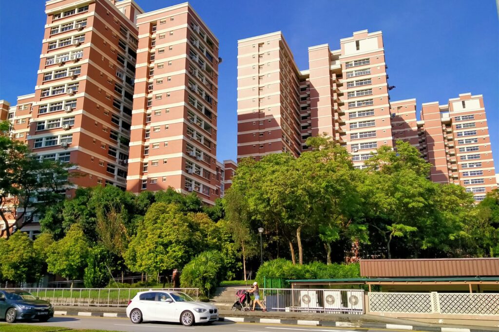 Tall, modern HDB apartment buildings in Singapore, featuring pink and orange tones, set against a bright blue sky with lush green foliage in the foreground.
