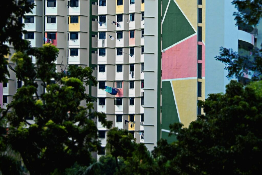 Framed by dark, leafy foliage in the foreground, high-rise apartment blocks rise up featuring distinctive green, pink, and yellow geometric murals on their facades. The dense grid of windows is punctuated by laundry hanging on poles to dry, adding a sense of lived-in reality to the colorful urban architecture.