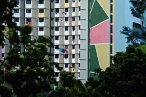 Framed by dark, leafy foliage in the foreground, high-rise apartment blocks rise up featuring distinctive green, pink, and yellow geometric murals on their facades. The dense grid of windows is punctuated by laundry hanging on poles to dry, adding a sense of lived-in reality to the colorful urban architecture.