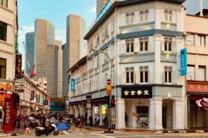 This vibrant street scene features a traditional white corner building with shuttered windows and Chinese signage, contrasting sharply with the towering modern skyscrapers in the background. Pedestrians and a scooter navigate the bustling intersection, which is lined with colorful shops and showcases a distinctive blend of historic charm and contemporary city life.
