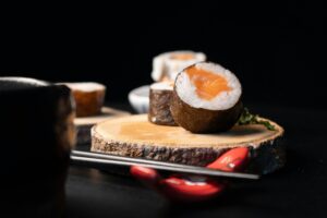 A close-up shot highlights a fresh salmon sushi roll resting on a rustic wooden slice against a dramatic dark background. In the foreground, black chopsticks sit on a glossy red rest, while additional blurred sushi pieces add depth to the composition.