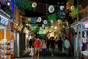 A bustling street market comes alive at night under a canopy of vibrant, multi-colored fairy lights and decorative hanging lanterns. Pedestrians, many wearing protective face masks, stroll past souvenir shops and stalls in what appears to be a festive Chinatown district.