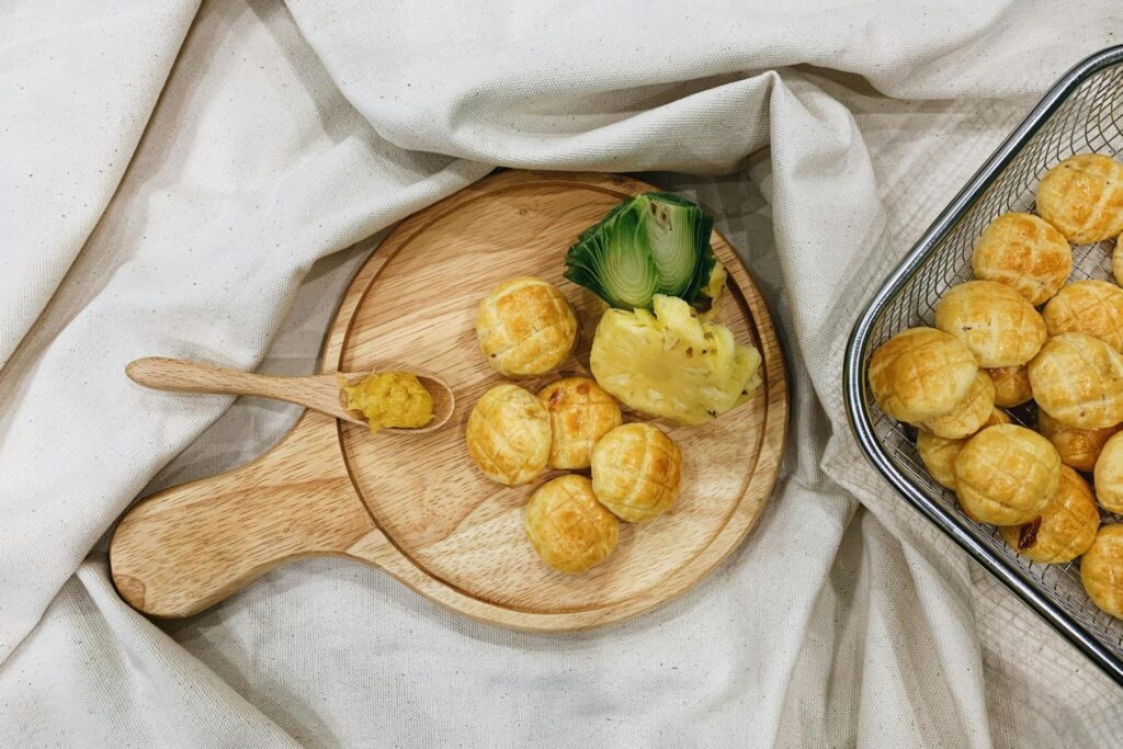A wooden serving board displays a cluster of golden-brown pineapple tarts alongside a slice of fresh pineapple and a small wooden spoon holding jam. To the right, a wire basket holds a large batch of the same glazed pastries, all resting on a draped, cream-colored fabric background.