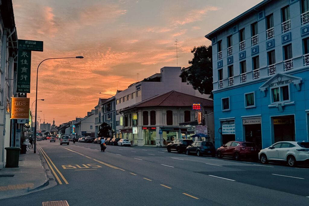 Singapore street at sunset with colorful sky, low‑rise shophouses, and light traffic along a quiet road.