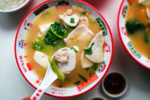 Top-down view of a colorful bowl of fish soup with slices of white fish, leafy greens, and herbs in a light broth. A hand holds a spoon lifting a piece of fish and vegetables from the bowl.