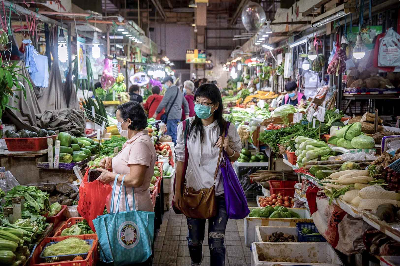 Busy wet market aisle lined with fresh vegetables, fruits, and produce, with shoppers walking and browsing under bright overhead lights.