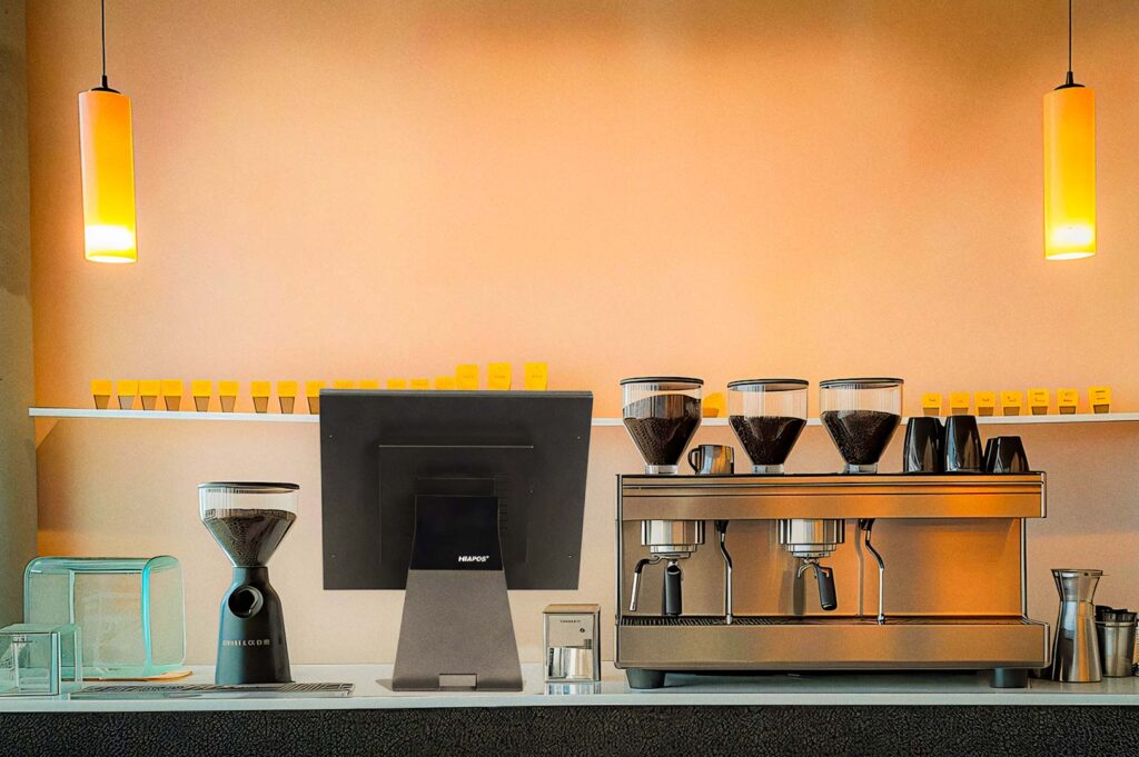Minimalist coffee bar with warm pendant lighting, featuring a commercial espresso machine, grinders, a touchscreen register, and neatly arranged espresso cups on a shelf.