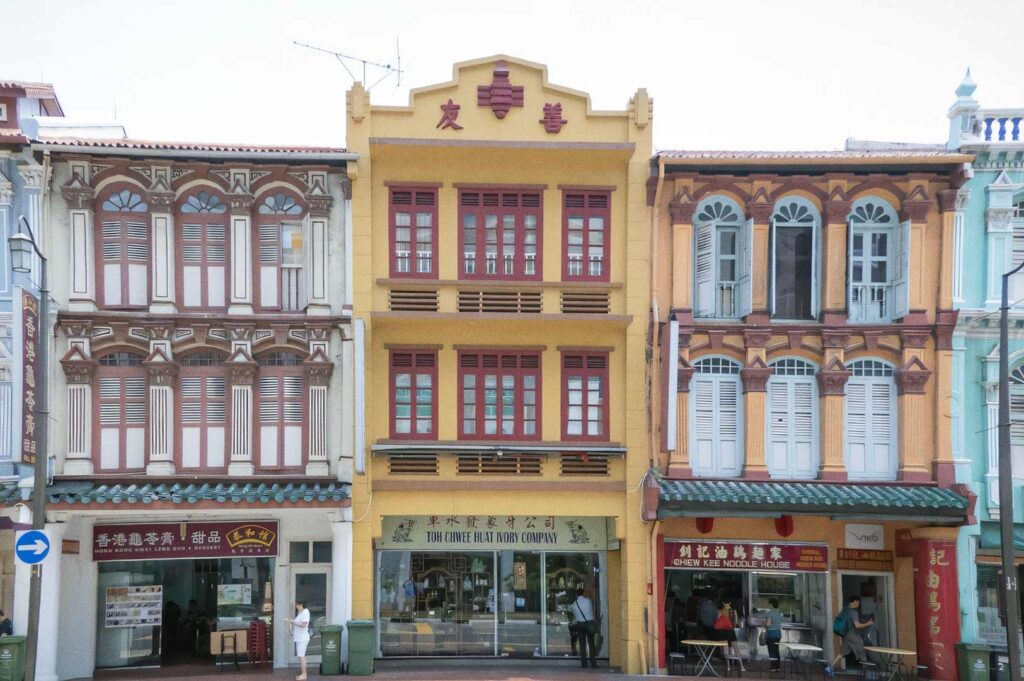 Row of colorful historic shophouses with ornate window shutters and decorative facades. The yellow central building has red windows and a shop on the ground floor, with neighboring pastel buildings featuring intricate architectural details.