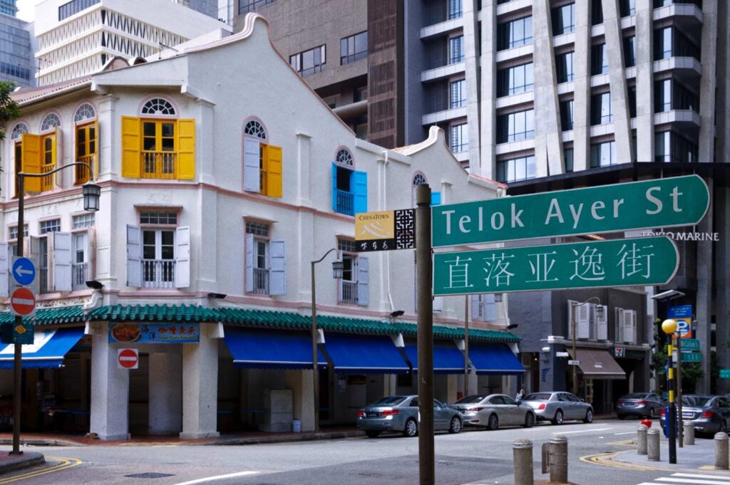 Street sign for Telok Ayer Street with Chinese text, shophouse with colorful shutters, and tall buildings behind.