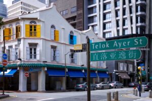 Street sign for Telok Ayer Street with Chinese text, shophouse with colorful shutters, and tall buildings behind.