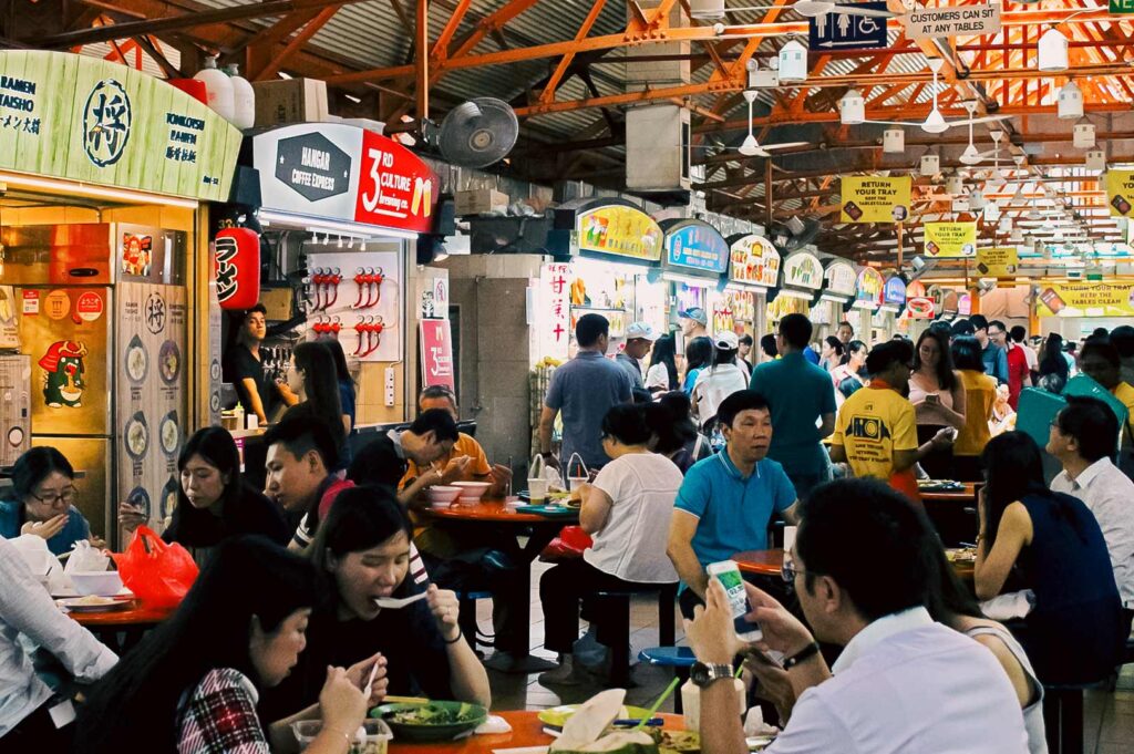 Crowded Singapore hawker centre with diners enjoying local dishes and food stalls lining the walkway.