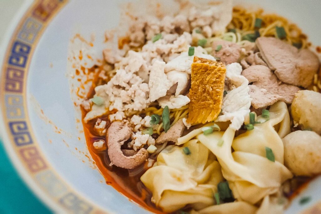 Close-up of Singapore Bak Chor Mee topped with minced pork, pork slices, wontons, and crispy lard in spicy chili sauce.