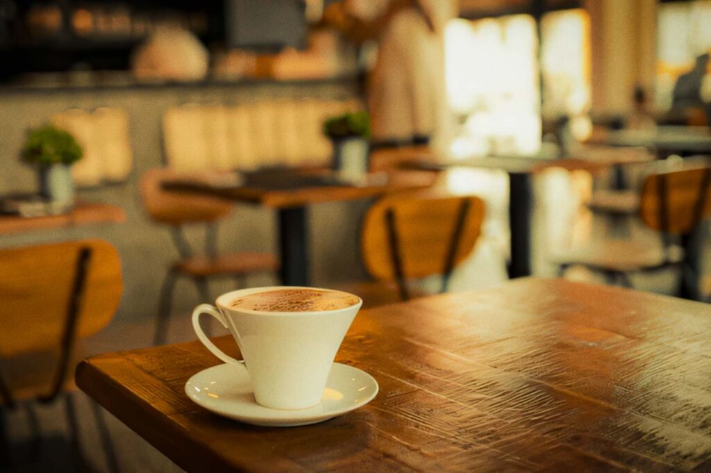White cup of coffee on a wooden table inside a cozy café, with soft warm lighting and blurred seating in the background.