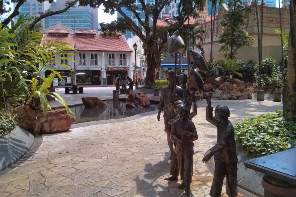 Bronze statue installation at Telok Ayer Singapore showing cultural procession with fish-shaped lanterns, tropical landscaping, colonial buildings, and modern high-rises.