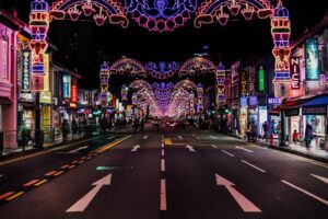 This image captures a vibrant street in Little India, Singapore, decorated with elaborate, glowing neon arches and festive light displays for Deepavali. The long exposure shot features a symmetrical view of the road, lined with brightly lit shops, colorful signs, and people strolling along the sidewalks under the purple and gold illuminations.