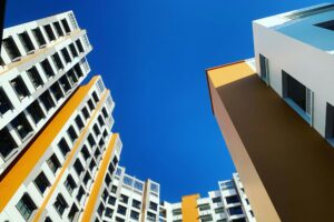 A worm's-eye view captures several modern high-rise buildings reaching toward a clear, vibrant blue sky. The architecture features a crisp white facade accented by bold orange sections and uniform rows of dark-framed windows.