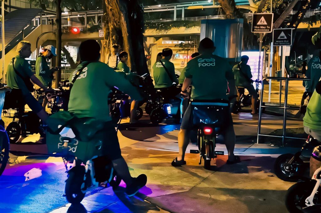 Group of delivery riders on e‑bikes gathered at night under city lights.