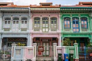 This image features a row of vibrant, pastel-colored shophouses with ornate architectural details, including decorative tiles and intricate window frames. A pink house in the center displays a "For Sale" sign on its front gate, while blue and green trash bins sit neatly along the sidewalk in front of the neighboring green unit.