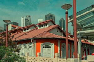 This vibrant image features a traditional orange building with a terracotta tiled roof, set against a backdrop of modern skyscrapers under a teal sky. The architecture showcases classic white-shuttered windows and ornate detailing, juxtaposed with contemporary street lamps and structural elements in the foreground.