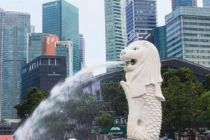 The image features the iconic Merlion statue in Singapore, a mythical creature with a lion's head and a fish's body, spouting a stream of water from its mouth. In the background, a dense cluster of modern skyscrapers bearing corporate logos like Deutsche Bank and Bank of America creates a striking urban skyline.