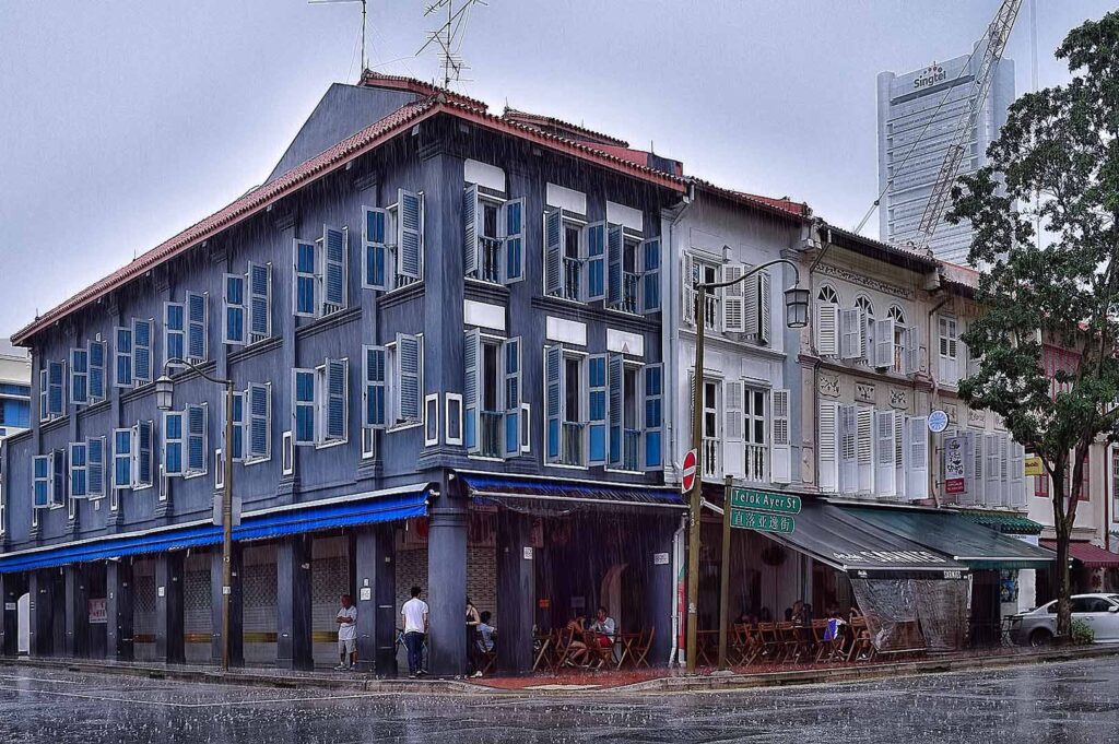Street sign for Telok Ayer Street with Chinese text, shophouse with colorful shutters, and tall buildings behind.