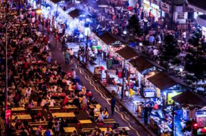 Aerial night view of a vibrant outdoor food street filled with dining tables, food stalls decorated with string lights, and crowds enjoying local street food.